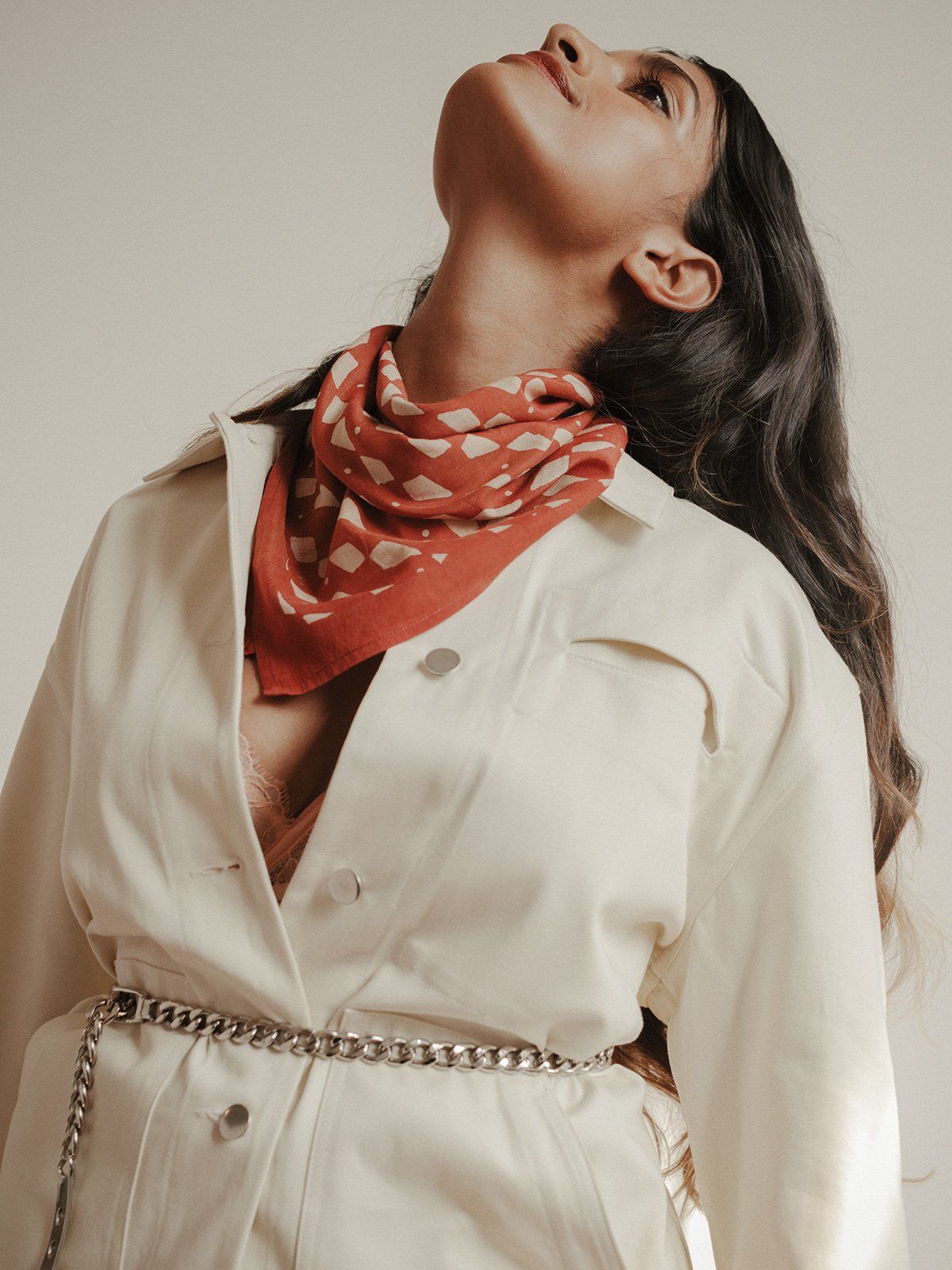 Deep earthy red block printed bandana scarf by SUNDAY/MONDAY. Worn around neck with a cream jacket for fall.